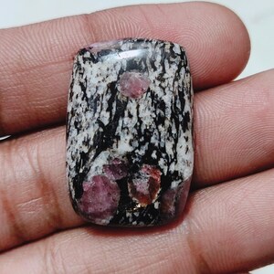 Könnte beinhalten: Ein rechteckiger Edelstein mit einem einzigartigen Muster aus schwarzen, weißen und rosa Farbtönen. Der Stein hat ein gesprenkeltes Aussehen mit mehreren prominenten rosa Einschlüssen. Die polierte Oberfläche reflektiert das Licht und unterstreicht die natürliche Schönheit des Steins.