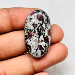 May include: An oval-shaped cabochon gemstone featuring a speckled white and black granite-like matrix embedded with several distinct, deep red, rounded inclusions distributed across the polished, smooth, and slightly domed surface.
