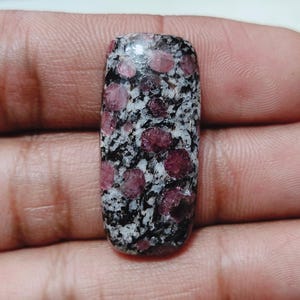 May include: A rectangular gemstone with a speckled pattern of black, white, and deep pink. The polished stone has a smooth surface and rounded edges. The gem is held in a hand.