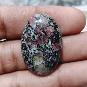 Peut inclure: Une pierre précieuse ovale avec une base mouchetée noire et blanche, ornée de grappes d'inclusions rose foncé et rouge. La surface polie reflète la lumière, mettant en valeur les motifs et les couleurs naturels de la pierre. Idéale pour la joaillerie.