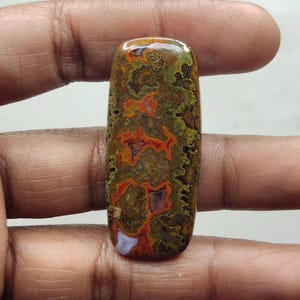 Puede incluir: Una gema rectangular pulida, con un patrón moteado de tonos marrones, verdes y naranjas. La piedra tiene un acabado brillante y extremos redondeados. La apariencia general es terrosa y natural, adecuada para joyería o fines decorativos.