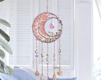 May include: A crescent moon-shaped wall hanging with pink and purple floral accents and dangling crystals. The moon is adorned with pink flowers and a central crystal pendant. Strands of beads and crystals hang below, with moon and star charms.