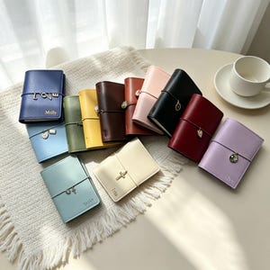 May include: A collection of small, rectangular, faux leather journals in various colors including blue, green, yellow, brown, pink, black, red, and lavender. Each journal features a thin elastic band with a metal charm and personalized text.