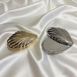 Op de afbeelding: Twee schelpvormige compacte spiegels. Eén is gesloten en goudkleurig, de andere is open en zilverkleurig, met de tekst "Lookin Good" op de spiegel. Beide liggen op een witte satijnen ondergrond.