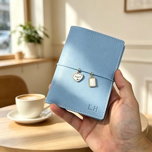 Puede incluir: Una funda de cuero azul claro para cuaderno con una banda elástica vertical y dos dijes colgantes: un dije en forma de corazón con la palabra amore y un dije rectangular blanco. Las iniciales L.H. están grabadas en la esquina inferior.