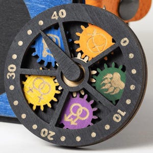 May include: A close-up of a wooden clock face with a black frame and a central hand. The clock features colorful gears with various symbols and numbers around the edge. The gears are blue, orange, green, yellow, and purple.