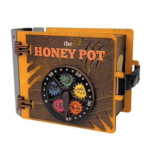 May include: A wooden box with a dark brown front panel featuring the text "the HONEY POT" in orange. A circular dial with a black pointer and colorful gear-shaped icons sits in the center. The box has light orange edges, a black strap, and hinges.