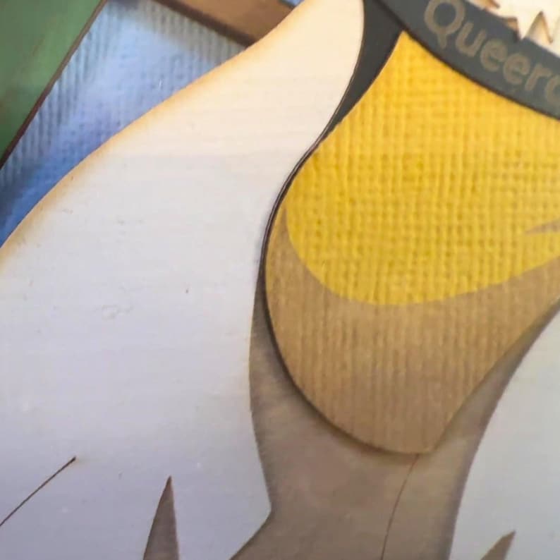 Puede incluir: Un recorte de madera en capas que presenta una superficie blanca lisa y una secci&oacute;n amarilla texturizada. En la parte superior se observa un segmento de borde curvo oscuro con el texto parcial "Queer" impreso en color claro sobre una base de madera.