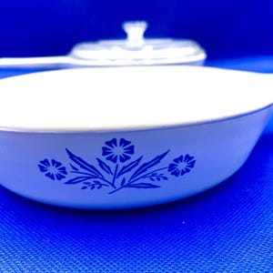 May include: White casserole dish with a blue floral design. The dish has a handle and a clear glass lid is visible in the background. The blue floral pattern is on the side of the dish.