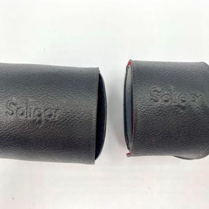 Puede incluir: Dos estuches de lente de cuero negro, cada uno con la palabra "Soligor" en relieve. Los estuches son cilíndricos, con una superficie texturizada. Un estuche está abierto, revelando un interior rojo.