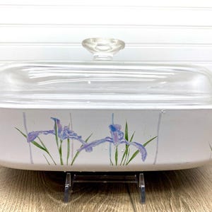 Puede incluir: Fuente rectangular blanca para horno con tapa de cristal transparente. El plato presenta un diseño floral de iris moradas y tallos verdes. La tapa tiene un pomo de cristal transparente. El plato está sobre un soporte de metal negro.