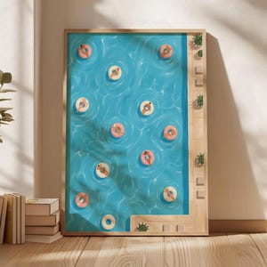 Op de afbeelding: Een ingelijste kunstdruk toont een zwembadscène. Het zwembad is gevuld met lichtblauw water en toont verschillende mensen die op roze en witte opblaasbare ringen drijven. Het kunstwerk is ingelijst in een licht houten frame.