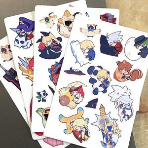 May include: Three white rectangular sticker sheets featuring an assortment of colorful, small, chibi-style anime character illustrations. The designs include various figures, creatures, and weapons in a cartoon aesthetic with bold outlines and vibrant colors.