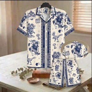 May include: A cream-coloured two-piece matching set, comprising a short-sleeved button-down shirt and shorts. The set features a blue cartoon print of characters and the text "DUNGEON CRAWLER CARL". The shirt has a collar and the shorts have a drawstring.
