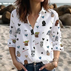 May include: White button-down shirt featuring a Star Wars character print. The long-sleeved shirt has a classic collar and is decorated with images of Darth Vader, Yoda, Stormtroopers, and other characters. The shirt is worn with blue jeans.