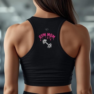 Pode incluir: Top desportivo preto com a inscrição "GYM MOM" em rosa gotejante e um gráfico de haltere prateado. O top tem um estilo nadador, perfeito para treinos. O fundo é um ginásio desfocado.