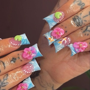 Può includere: Unghie lunghe scolpite con un design unico. Ogni unghia presenta un effetto marmorizzato blu chiaro e bianco, adornato con fiori rosa 3D e piccoli dettagli dorati. Alcune unghie hanno un design verde arrotondato.