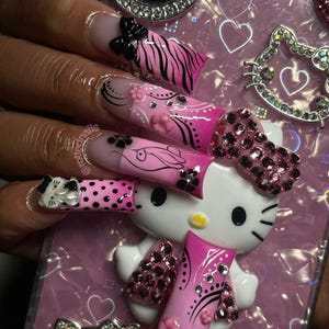 May include: Long, embellished acrylic nails in shades of pink, black, and white. Designs include zebra stripes, polka dots, floral accents, and a 3D Hello Kitty figure. The nails are adorned with rhinestones and bows.