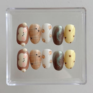 May include: A set of ten artificial nails in a clear plastic case. The nails feature various designs, including a brown bunny, plaid, pumpkins, a swirl of green and brown, and yellow with small brown hearts.