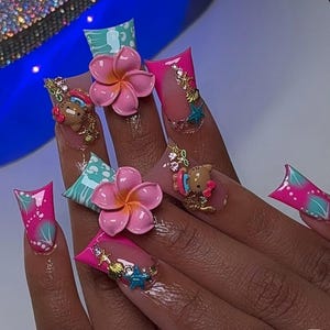 May include: A set of long, square-shaped artificial nails. The nails are painted with a gradient of pink and clear polish, adorned with 3D pink flowers, gold embellishments, and cartoon character accents. The design includes teal and pink accents.