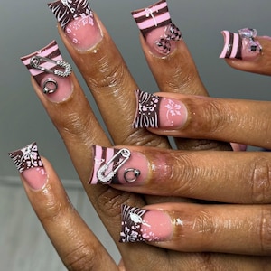 May include: A set of long, square-shaped artificial nails. The nails feature a combination of designs, including pink and brown stripes, floral patterns, and silver safety pin and ring accents. Some nails have clear bow-shaped embellishments.