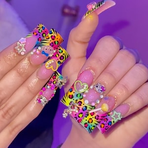 Pode incluir: Unhas acrílicas longas e coloridas com um design de estampa de leopardo arco-íris. Cada unha é adornada com strass, berloques e glitter, incluindo corações, estrelas e um pirulito. As unhas têm uma base rosa e uma ponta francesa.