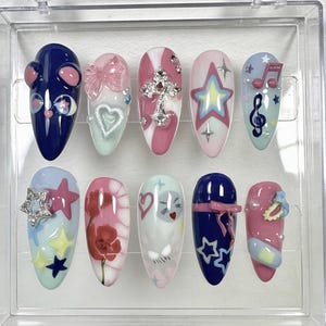 May include: A set of ten artificial nails with various designs. The nails feature a range of colors, including blue, pink, and white, and are adorned with embellishments such as bows, stars, and crosses. Some nails have painted designs, including flowers and musical notes.