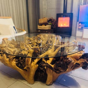 May include: A unique coffee table featuring a natural wood base with a clear glass top. The base is made of intricately shaped, light brown wood, showcasing the natural grain and texture. The table is a statement piece for any living room.