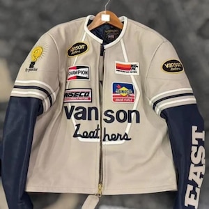 May include: A beige leather jacket with navy blue sleeves and white accents. The jacket features multiple patches with text and logos, including "Vanson Leathers" and "EAST" on the sleeve. A zipper runs down the front.