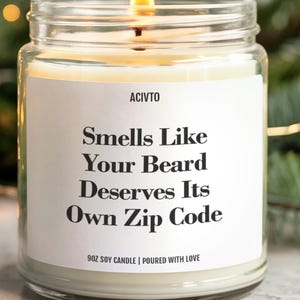 Peut inclure: Une bougie de soja allumée dans un bocal en verre transparent. L'étiquette indique "Smells Like Your Beard Deserves Its Own Zip Code". La bougie est de 255g et coulée avec amour. L'arrière-plan comprend des branches de sapin et des guirlandes lumineuses.