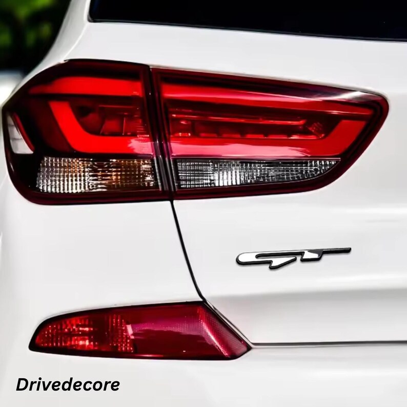 May include: Rear view of a white car featuring a red tail light and a black "GT" emblem. The tail light has a modern design. The text "Drivedecore" is visible at the bottom of the image.