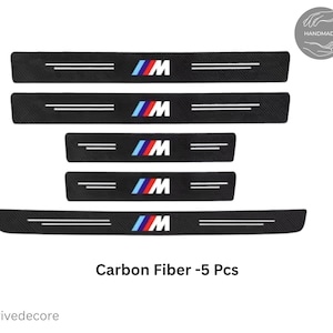 Puede incluir: Juego de cinco protectores de umbral de puerta de coche de fibra de carbono negra. Cada pieza presenta el logotipo "M" en azul, rojo y blanco, con dos líneas blancas paralelas. El texto "Carbon Fiber -5 Pcs" se muestra debajo.