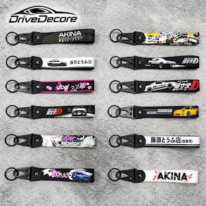 May include: A collection of twelve fabric keychains featuring black metal clips and rings. Each strap displays unique automotive-themed graphics, including Japanese text, car illustrations, and logos like Akina, Initial D, and Midnight.