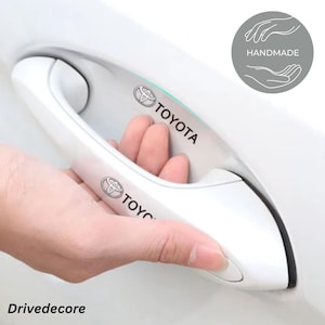 Könnte beinhalten: Weißer Autotürgriff mit dem schwarzen Toyota-Logo. Eine Hand öffnet den Griff. Das Wort "HANDMADE" steht in einem grauen Kreis oben rechts. Das Wort "Drivedecore" steht unten links.