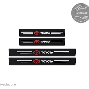 May include: Four black car door sill protectors with the Toyota logo in red and the word "TOYOTA" in white. Each protector has two white horizontal lines. The upper right corner has a gray circle with the word "HANDMADE".
