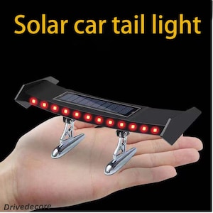 Op de afbeelding: Zwart zonne-auto-achterlicht met rode LED-verlichting en een zonnepaneel. De tekst "Solar car tail light" wordt in het geel weergegeven. Het achterlicht wordt in een hand gehouden.