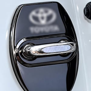 May include: A glossy black car door handle cover featuring a centered horizontal metallic handle and a blurred Toyota logo at the top. The accessory is mounted on a white vehicle surface, with the DriveDecore brand name visible at the top left.