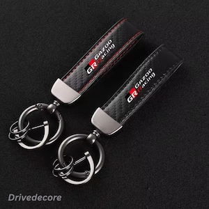 May include: Two black keychains featuring a carbon fibre texture and red stitching. Each keychain has a silver metal clasp and a round ring, with the Gazoo Racing GR logo printed in white and red on the front of the black strap material.