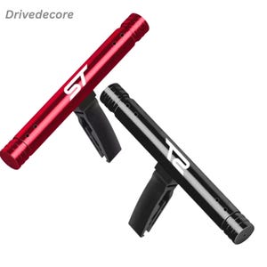 Pode incluir: Dois acessórios para ventilação de carro, um vermelho e outro preto. Cada um tem as letras "ST" impressas em branco. Os acessórios são cilíndricos com vários orifícios pequenos e um clip preto para fixação. A marca "Drivedecore" está no canto superior esquerdo.