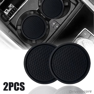 May include: Two black car cup coasters with a textured honeycomb pattern. The coasters are round and designed to fit in a car's cup holders. The image also shows the car's interior, including the cup holder area.