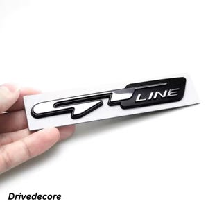 May include: A black and silver GT Line emblem on a white rectangular backing. The emblem features the text "GT LINE" in a bold, sans-serif font. The emblem is held by a hand against a white background.