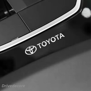 Könnte beinhalten: Ein glänzend schwarzes Interieur-Zierteil für Fahrzeuge mit dem silbernen Toyota-Logo und dem Schriftzug TOYOTA in einer serifenlosen Schriftart. Ein dünner, metallisch silberner Zierstreifen verläuft diagonal über den oberen Teil.