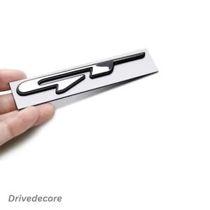 May include: A silver and black GT emblem. The emblem is rectangular with a glossy black outline and a silver interior. The letters "GT" are stylized and integrated into the design. The text "Drivedecore" is at the bottom.