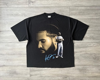 May include: Black t-shirt featuring a portrait of a man with a beard and a smaller image of a man in a white outfit. The portrait is in shades of gold and brown, with a blue signature at the bottom. The t-shirt is made of a soft material.