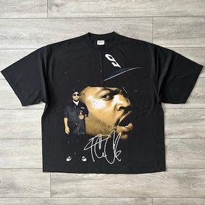 May include: A black short-sleeve t-shirt featuring a large graphic print of Ice Cube. The design includes a full-body image of the artist, a prominent close-up of his face wearing a baseball cap, and a white stylized signature printed below.