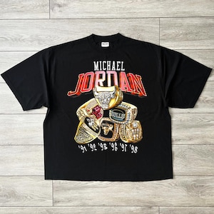 May include: Black t-shirt with the name "MICHAEL JORDAN" in white and red lettering above a graphic of championship rings. The rings are gold with various gemstones and the years '91, '92, '93, '96, '97, and '98.
