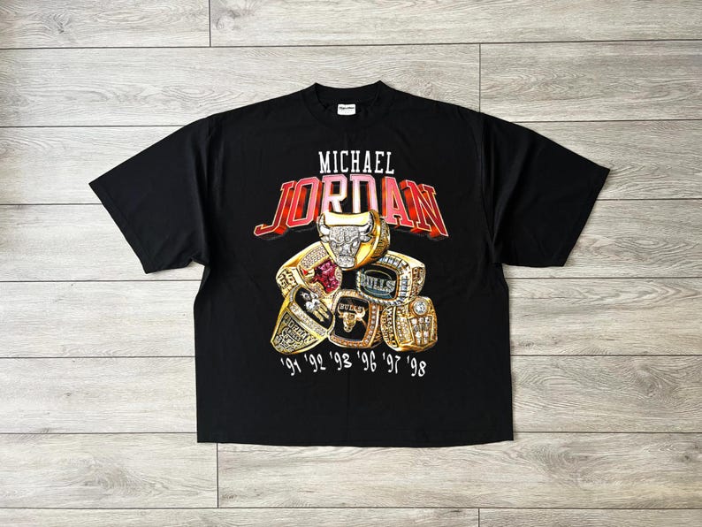 May include: Black t-shirt with the name "MICHAEL JORDAN" in white and red lettering above a graphic of championship rings. The rings are gold with various gemstones and the years '91, '92, '93, '96, '97, and '98.