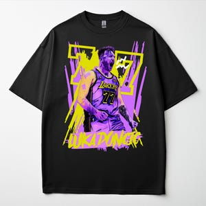 Puede incluir: Camiseta negra con un gráfico de un jugador de baloncesto con la camiseta de los Lakers, número 77. El diseño utiliza acentos amarillos y morados, con el nombre del jugador, LUKA DONCIC, impreso en amarillo.