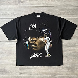 May include: A black short-sleeve t-shirt featuring a large graphic print of a baseball player wearing a New York Yankees helmet. The design includes a close-up portrait of the player, a full-body image of the player in a uniform, and a white signature.