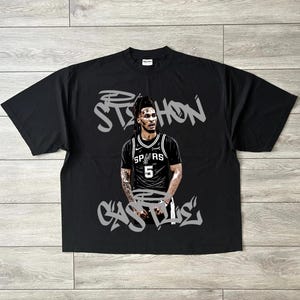 May include: Black t-shirt featuring a graphic of a basketball player in a Spurs jersey. The player's name, "Stephon Castle," is written in stylized gray graffiti font above and below the image. The shirt is a crew neck style.